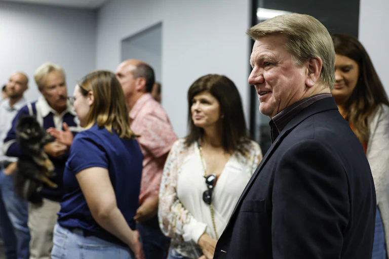 A white man with blonde hair stands in in a room with several people in the background.