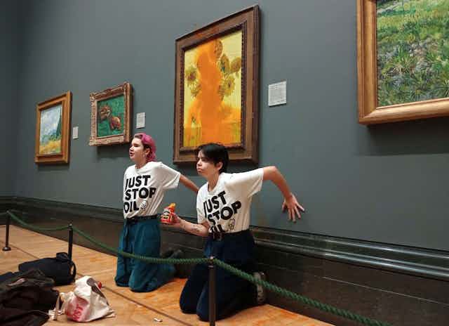 Protesters sit in front of defaced painting with hands glued to wall