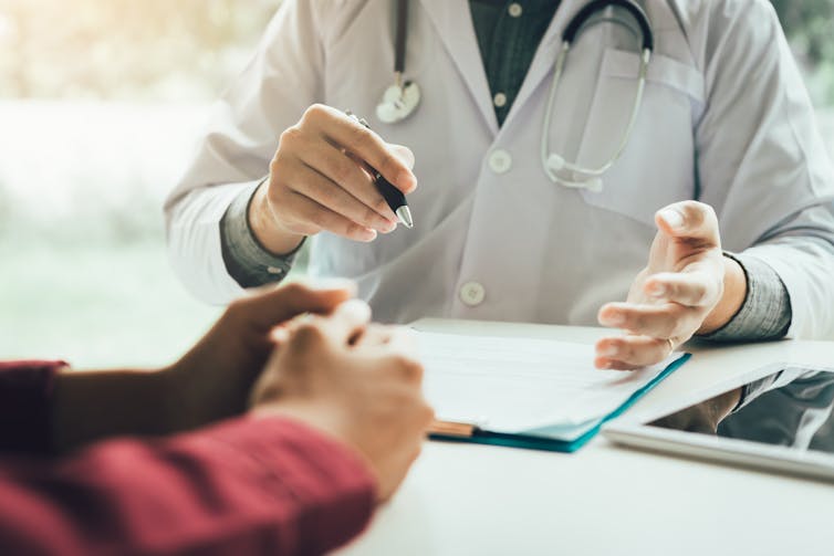 A doctor speaking with a patient in their office. The doctor has a clipboard in front of them on their desk.