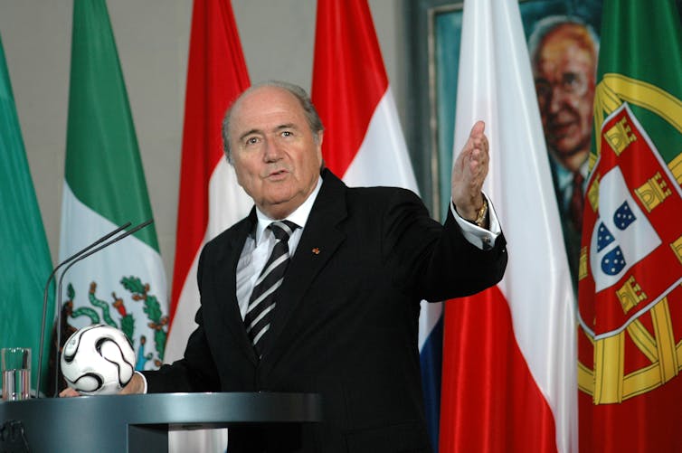 A man gesticulates while speaking at a lectern with a small football on it.