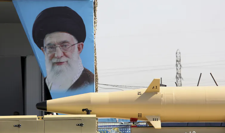 A portrait of a bearded man with a turban is seen behind a large missile.