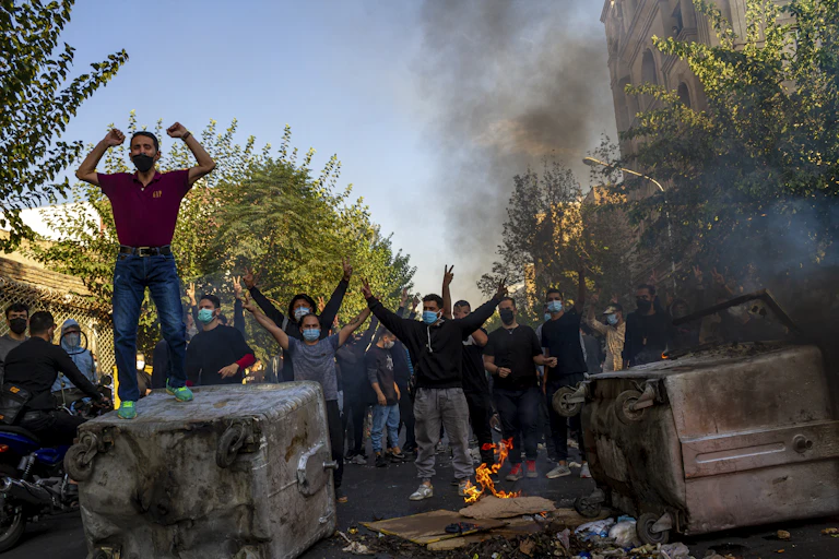 Small fires burn as protesters, mostly men, stand with raised first on overturned vehicles.