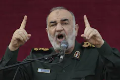 A man with a short beard in a military uniform gestures with both hands as he speaks.