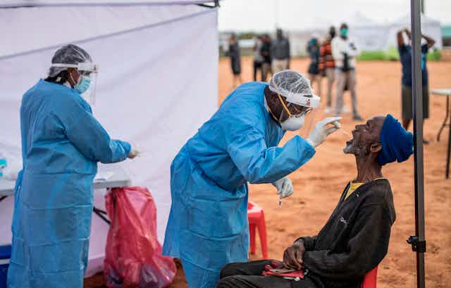 Dos sanitarios con bata toman una muestra de la garganta de un hombre bajo una tienda de campaña en un paisaje seco de África