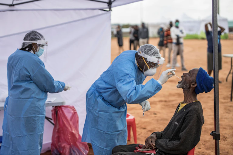 Dos sanitarios con bata toman una muestra de la garganta de un hombre bajo una tienda de campaña en un paisaje seco de África