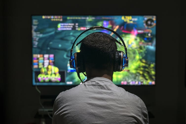 A teenager is seen from behind, playing online, with a headset on
