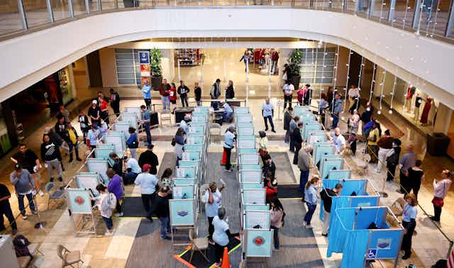 A view from above shows many small cubicles in which people are standing and making selections on screens.