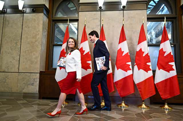 une femme et un homme qui tiennent des feuillets marchent devant des drapeaux du Canada