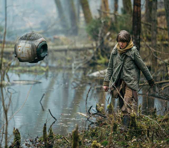A small robot seen hovering over a wetland-type landscape and a teen is walking in a military-style coat.
