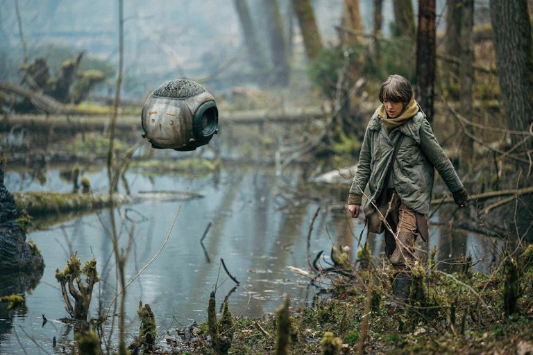 A small robot seen hovering over a wetland-type landscape and a teen is walking in a military-style coat.