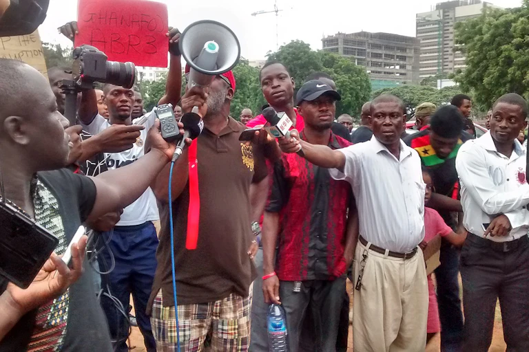 Man using a megaphone in front of a crowd of people as men with cameras and microphones record him.
