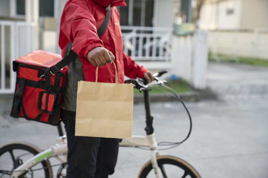 A person holding out a brown bag while holding a bicycle, and with a courier bag on his back.