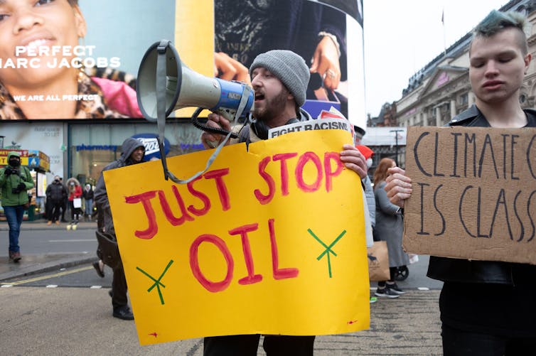 Um jovem fala através de um megafone enquanto segura uma placa amarela onde se lê Just Stop Oil em uma demonstração no Picadilly Circus, em Londres