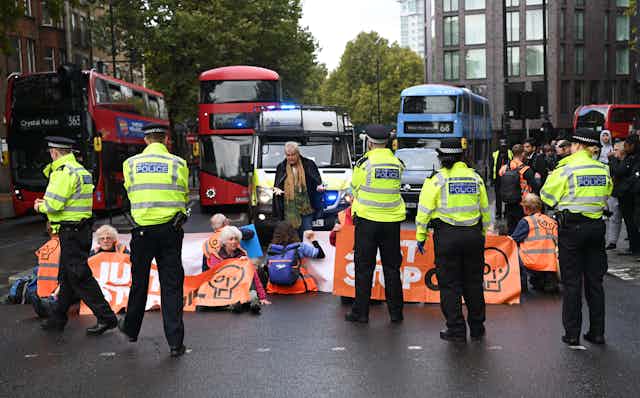 Police approach protesters siting in a road with orange Just Stop Oil banners, blocking buses and police vehicles