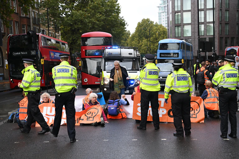 Police approach protesters siting in a road with orange Just Stop Oil banners, blocking buses and police vehicles