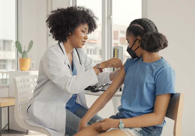 A woman in a white coat with a stethoscope putting a bandage on the arm of a girl in a blue T-shirt and black face mask