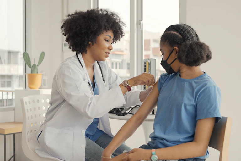 A woman in a white coat with a stethoscope putting a bandage on the arm of a girl in a blue T-shirt and black face mask