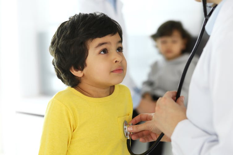 A child in a yellow T-shirt looking up at a health-care provider out of frame, who is examining the boy with a stethoscope