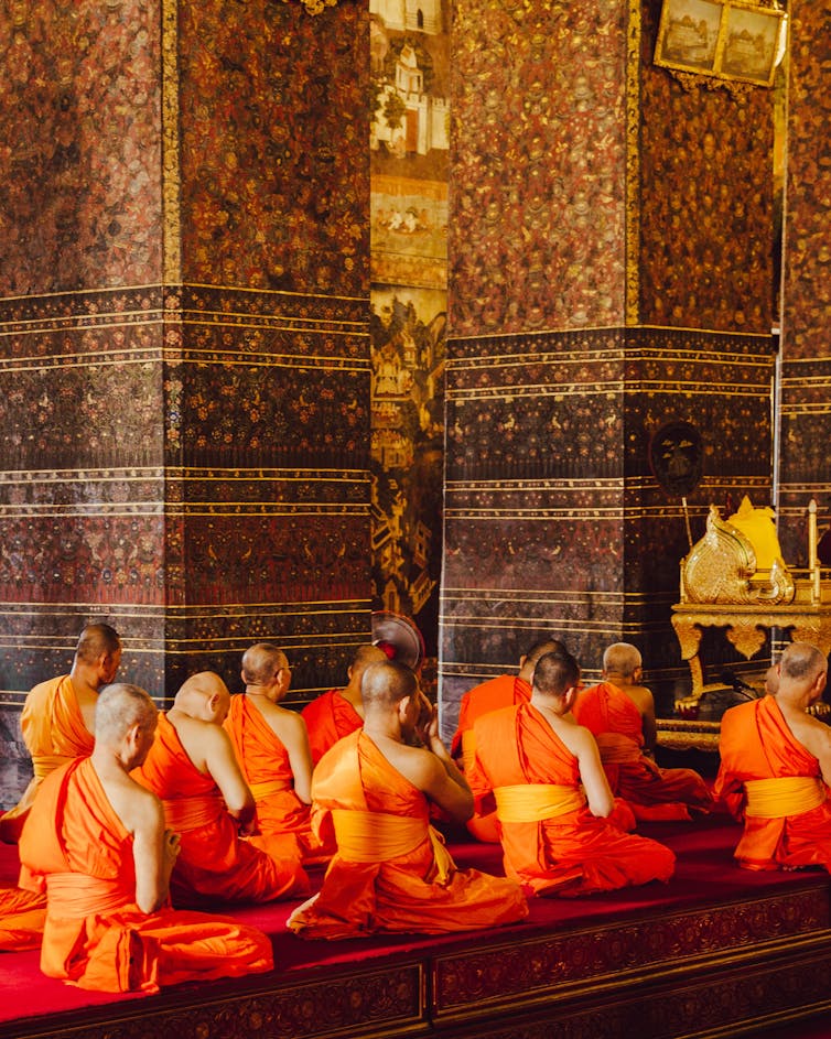 Buddhist monks with shaved heads and wearing traditional orange robes sit on the floor of a temple.