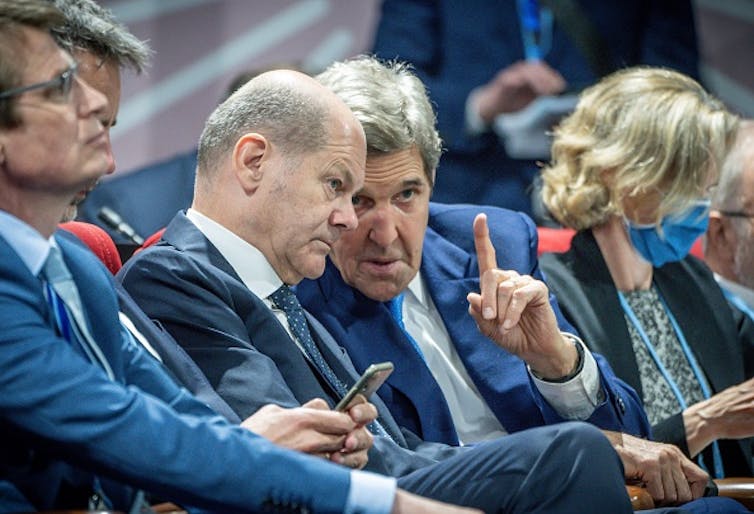 Kerry gestures with one hand as he speaks with Scholz amid other seated people.