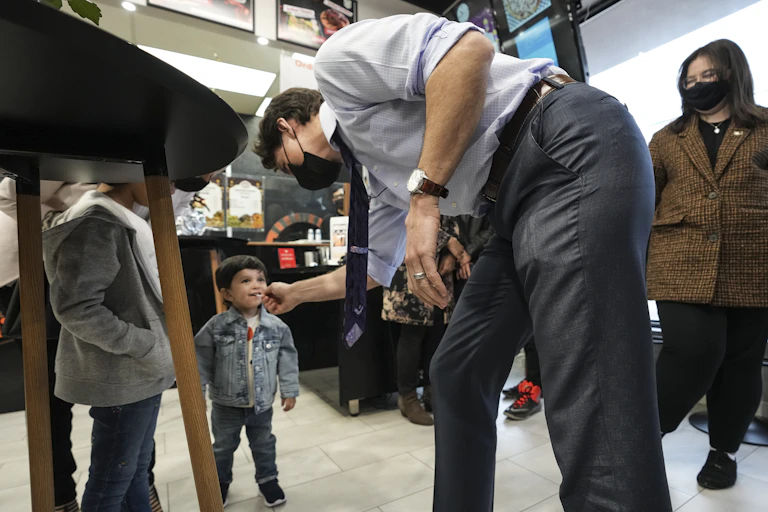 A dark-haired man wearing a face mask leans toward a smiling toddler who has a lollipop in his mouth.