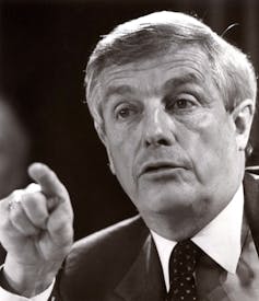A black-and-white photo shows a grey-haired man pointing his finger while talking.
