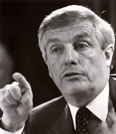 A black-and-white photo shows a grey-haired man pointing his finger while talking.