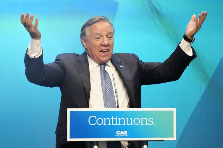 A man stands with raised hands behind a sign that reads Continuons.