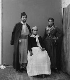A black and white photo shows a man and woman standing while another woman sits between them. They all wear long robes or skirts and have their heads covered.
