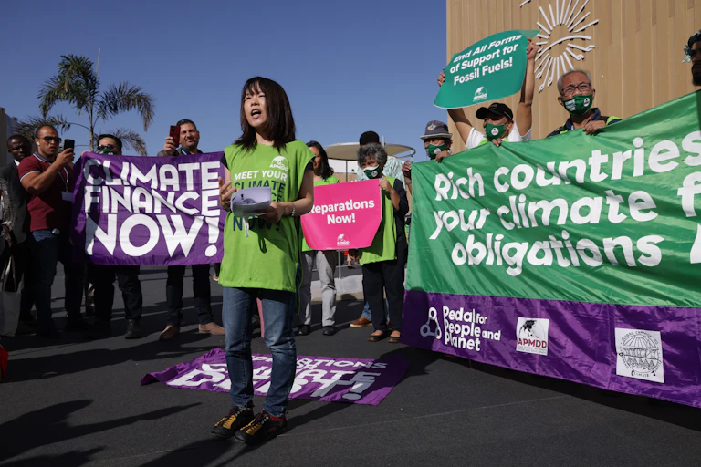 An activist stands with colleagues holding signs that read 'Climate finance now!' and 'Rich countries: pay your climate finance obligations now'.