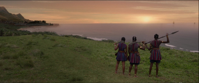 Three women in traditional uniform stand staring out at the ocean with shops approaching in the distance and a town nestled in a bay. They hold weapons and have their backs to the camera.