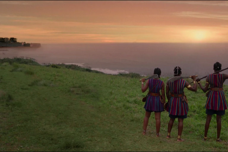 Three women in traditional uniform stand staring out at the ocean with shops approaching in the distance and a town nestled in a bay. They hold weapons and have their backs to the camera.