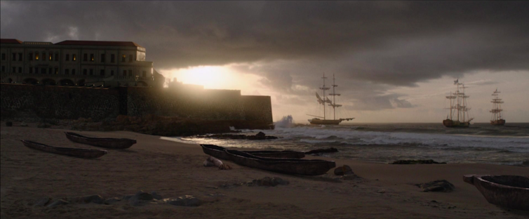 A moody still of an old port with ships approaching in a rough sea and the sun breaking through the clouds.