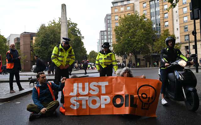 Two police officers stand over protesters seated behind an orange 'Just Stop Oil' banner in a road.