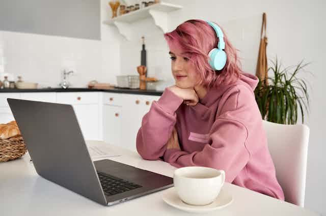 A young woman sits in front of her laptop in the kitchen, with headphones on.