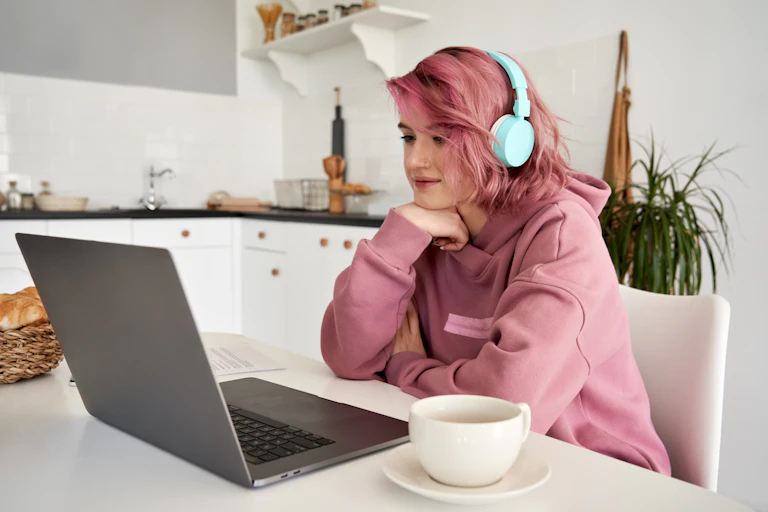 A young woman sits in front of her laptop in the kitchen, with headphones on.