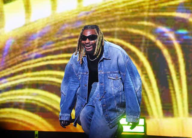 A man with golden dreads and sunglasses crouches on a stage with bright lights behind him. He smiles, revealing a gold grille.