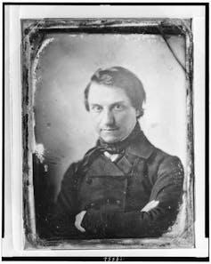 A black and white photo of a dark-haired man with his arms crossed.