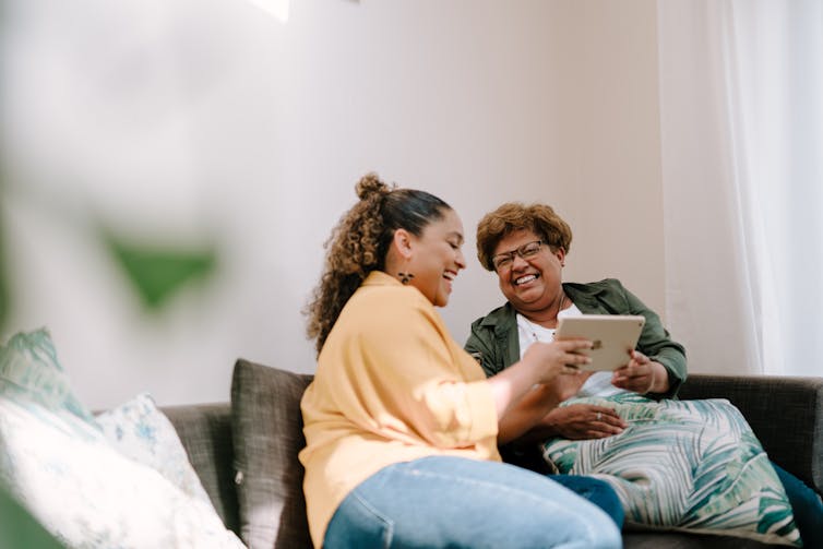 two women, one older and one younger, sit on a sofa - the older woman is holding a tablet in her hand