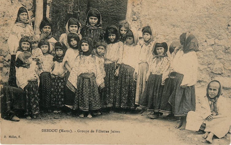 An old, black and white postcard shows a group of girls standing outside a doorway in skirts and kerchiefs.