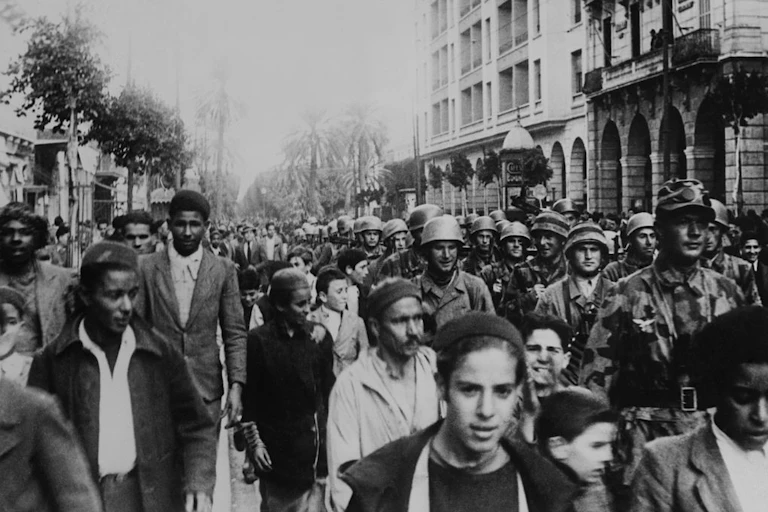 A black and white photo shows troops marching along a city street with palm trees as civilians walk alongside them.
