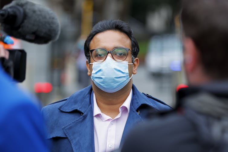 Man wearing mask being interviewed by press.