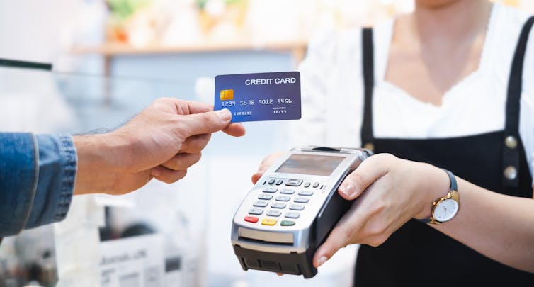 A hand holding a credit card reaching out toward a credit card reader machine, which is held by a worker wearing an apron