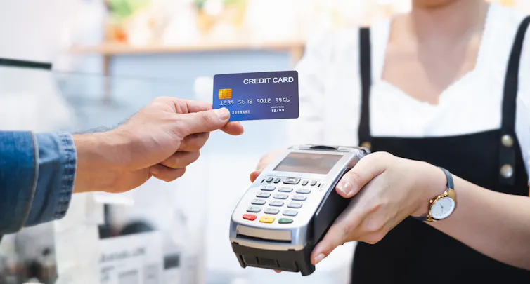 A hand holding a credit card reaching out toward a credit card reader machine, which is held by a worker wearing an apron