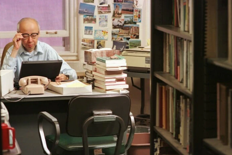An elderyl Asian man wearing glasses sits at a desk.