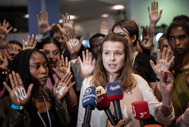 A young woman speaks at microphone and young people around her hold up their hands with the words