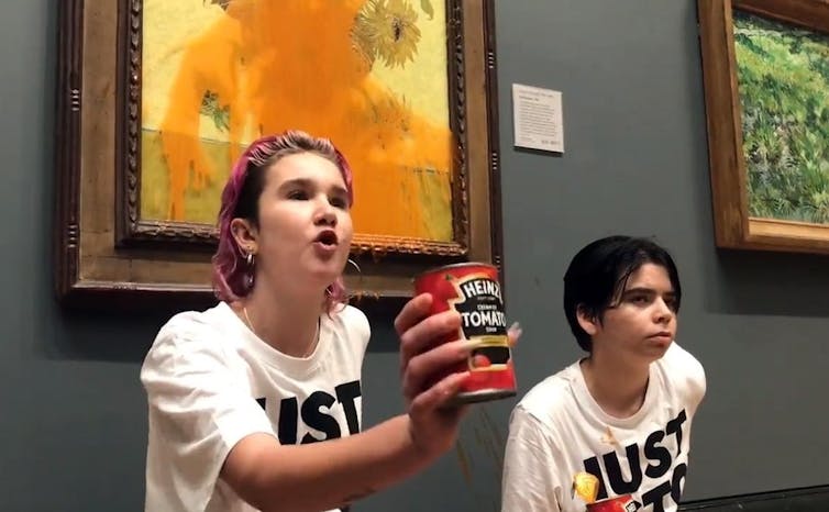 Protesters wearing 'Just Stop Oil' T-shirts stand next to Van Gogh's 'Sunflowers' painting, which has tomato soup streaming down the glass cover. One protester holds up the soup can for cameras. Both have one hand glued to the wall behind them.