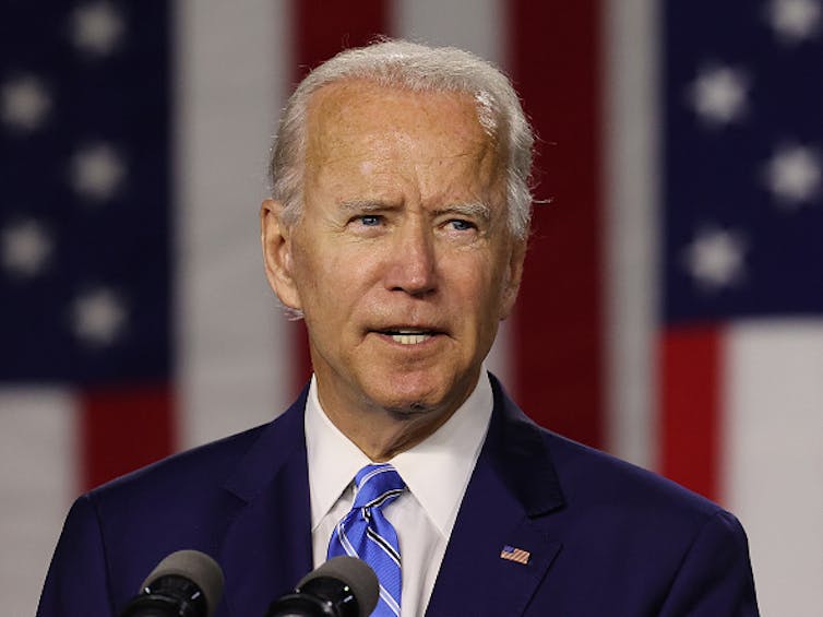 A man wearing a suit, shirt and tie is shown speaking into a microphone.