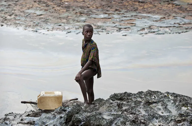 A boy stands on an oil-coated river bank.