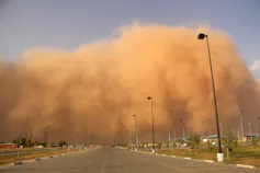 A large cloud of dust moving down a road against a clear blue sky.